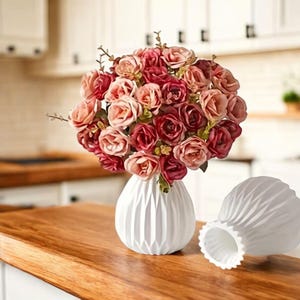 Peut inclure: Un bouquet de roses artificielles dans des tons de rose, mauve et rouge foncé disposé dans un vase blanc rond à côtes verticales. À côté du vase rempli se trouve un vase blanc identique vide avec un bord festonné et une surface plissée.