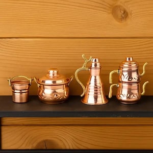 May include: A set of four miniature copper kitchen items including a small bucket with a handle, a lidded pot with side handles, a tall pitcher with a decorative gold-toned handle, and a stacked two-piece teapot with gold-toned handles.