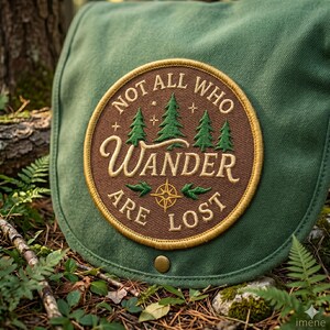 May include: A green canvas bag with a circular patch. The patch features the text "NOT ALL WHO WANDER ARE LOST" in a brown and gold design, with illustrations of trees and a compass. The bag is secured with a brass-colored button.