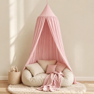 May include: A pink and white striped fabric canopy suspended from above, featuring a conical top and long, flowing panels. Beneath the canopy sits a beige floor cushion with several square pillows in beige and pink, plus a soft pink throw blanket.