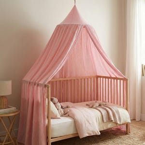 May include: A light pink fabric canopy hangs from the ceiling over a wooden crib. The canopy features a conical top and long, flowing panels that drape down to the floor. Inside the crib, there is a light pink blanket and two plush toys.