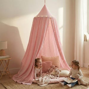 May include: A soft pink fabric canopy hanging from the ceiling, creating a conical tent shape. The canopy features draped fabric panels and is decorated with a string of small warm white lights. A circular floor mat sits underneath the canopy.