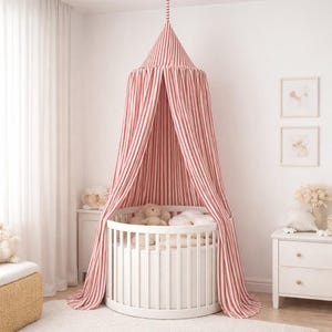 May include: A round white crib featuring a conical fabric canopy with vertical red and white stripes. The canopy hangs from the ceiling, draping over the crib, which contains several plush toys and soft white bedding inside the slatted frame.
