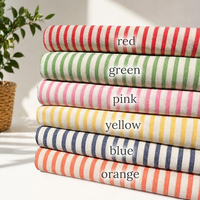 May include: A stack of six folded fabric bolts featuring vertical stripes in red, green, pink, yellow, blue, and orange. Each fabric bolt has the corresponding color name printed in black text on the front of the folded material.