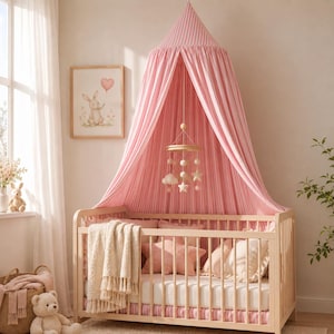 May include: A light pink and white striped fabric canopy hangs over a wooden crib. Inside the crib are pillows, a white blanket with tassels, and a hanging mobile with stars and clouds. A teddy bear sits on the floor next to a woven basket.