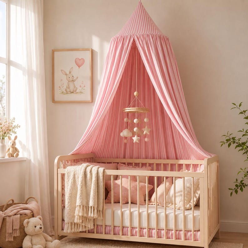 May include: A light pink and white striped fabric canopy hangs over a wooden crib. Inside the crib are pillows, a white blanket with tassels, and a hanging mobile with stars and clouds. A teddy bear sits on the floor next to a woven basket.