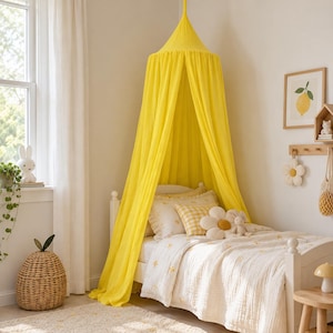 May include: A bright yellow fabric bed canopy suspended from the ceiling, draping over a white bed frame. The bed features a white quilt with small yellow floral patterns, a yellow and white checkered pillow, and a large white flower-shaped pillow.