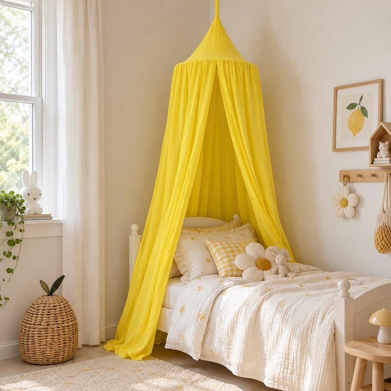 May include: A bright yellow fabric bed canopy suspended from the ceiling, draping over a white bed frame. The bed features a white quilt with small yellow floral patterns, a yellow and white checkered pillow, and a large white flower-shaped pillow.