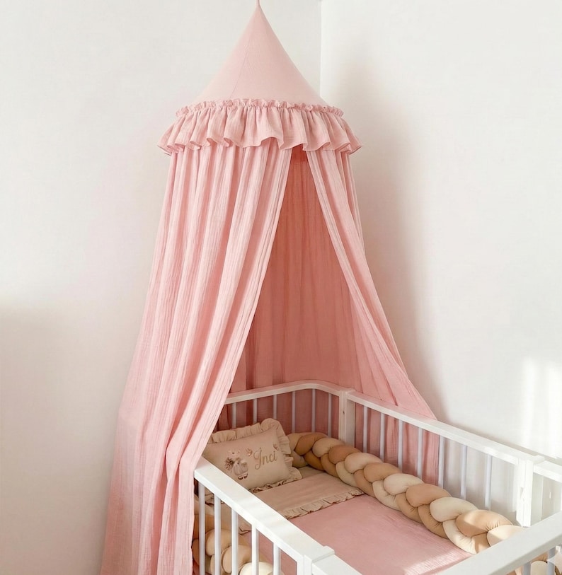 May include: A dusty pink fabric bed canopy with a ruffled top edge hanging over a white crib. Inside the crib is a rectangular pillow with the name Inci embroidered on it and a long, braided bumper in shades of beige, cream, and light pink.
