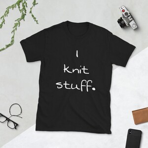 May include: A black short-sleeved t-shirt featuring the white text "I knit stuff." printed in a simple, handwritten-style font across the center of the chest. The garment has a classic crew neckline and a standard, straight-cut silhouette.