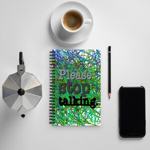 May include: A spiral-bound notebook with the text "Please stop talking" in gray and black. The cover features a colorful abstract design of blue, green, and red lines. A cup of coffee, a pencil, a phone, and a stovetop espresso maker are also visible.