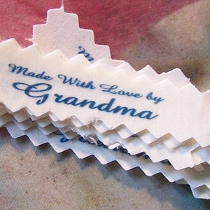 Puede incluir: Una pila de etiquetas de tela blanca con un borde festoneado. Las etiquetas est&aacute;n impresas con tinta azul y dicen "Made With Love by Grandma".