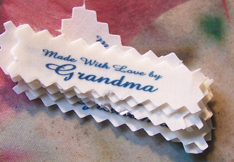 Puede incluir: Una pila de etiquetas de tela blanca con un borde festoneado. Las etiquetas est&aacute;n impresas con tinta azul y dicen "Made With Love by Grandma".
