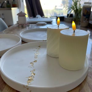 Puede incluir: Dos velas blancas en forma de pilar con llamas amarillas parpadeantes sobre una bandeja decorativa blanca. La bandeja tiene un diseño de hoja de oro y está rodeada de otras bandejas blancas. Las velas están sobre una mesa de madera.