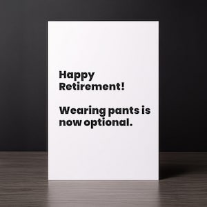 May include: A white greeting card with the text "Happy Retirement! Wearing pants is now optional." in black font. The card is on a wooden surface with a dark background.