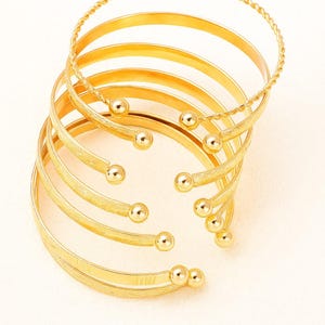 Peut inclure: Bracelets manchettes dorés. Les bracelets présentent un design en spirale avec plusieurs bandes, chacune ornée de petits accents ronds. Un bracelet a un design en corde torsadée. Les bijoux sont présentés sur fond blanc.