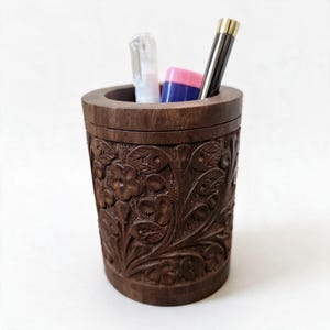 Peut inclure: Un porte-crayon cylindrique en bois brun foncé orné de motifs floraux et de feuilles sculptés à la main sur toute sa surface extérieure. Le récipient contient un stylo en plastique transparent et un stylo en métal noir et argenté avec un accent rose.