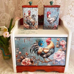 May include: White wooden bread box with a rooster and floral design. The box has two smaller containers with rooster and floral designs on top.