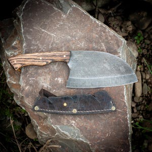 May include: A large, heavy-duty cleaver with a wooden handle and a wide, curved blade. The blade is gray and appears to be forged steel. A black leather sheath with the text "MANGILLO SMITHY" is below the cleaver. The cleaver is resting on a large, gray rock.
