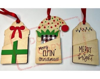 May include: Three wooden Christmas gift card holders. The first is a brown gift box with a green bow and the text "Merry Elfin' Christmas". The second is a brown elf hat with a green brim and the text "Merry Elfin' Christmas". The third is a brown gift tag with the text "Merry & Bright".