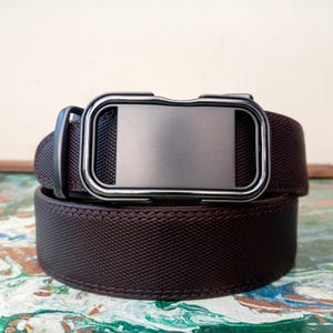 Op de afbeelding: Een bruine riem met een getextureerd oppervlak en een zilverkleurige rechthoekige gesp. De riem is opgerold, wat het ontwerp en de constructie laat zien. De gesp heeft een strakke, moderne uitstraling.