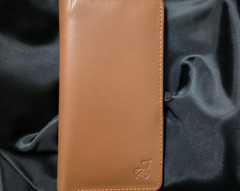 May include: A rectangular, tan leather wallet with a stitched border. The wallet has a small embossed design in the lower right corner. The background is a dark, textured fabric.