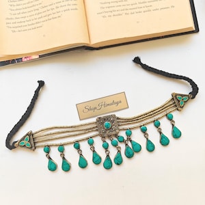 May include: A handcrafted choker necklace with turquoise teardrop pendants and a central square pendant. The necklace features a black cord and gold-toned metal accents, with the text "Shop Himalaya" on a small tag.
