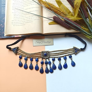 May include: A blue and gold choker necklace with teardrop-shaped pendants. The necklace has a square centre piece and triangular accents. The text "Shop Himalaya" is visible.