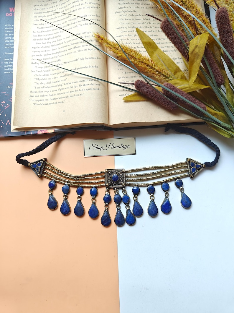 May include: A blue and gold choker necklace with teardrop-shaped pendants. The necklace has a square centre piece and triangular accents. The text "Shop Himalaya" is visible.