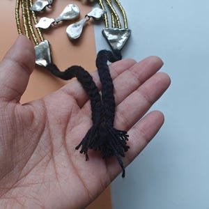 May include: A handcrafted necklace with silver-toned metal pendants and gold-coloured beaded strands. The necklace has a black braided cord with a tassel. The design includes geometric and organic shapes, creating a unique accessory.