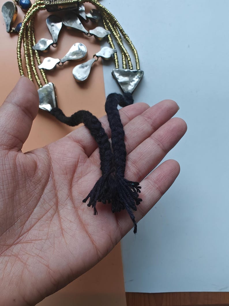 May include: A handcrafted necklace with silver-toned metal pendants and gold-coloured beaded strands. The necklace has a black braided cord with a tassel. The design includes geometric and organic shapes, creating a unique accessory.