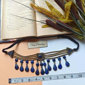 May include: A choker necklace with a black cord, gold-coloured metal details, and blue teardrop pendants. The necklace is about 25 cm long, with a central square and triangular side elements. The text "Shop Himalaya" is visible.