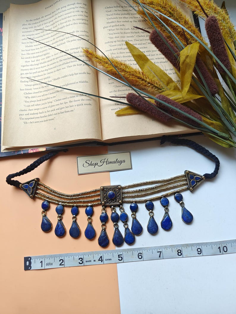 May include: A choker necklace with a black cord, gold-coloured metal details, and blue teardrop pendants. The necklace is about 25 cm long, with a central square and triangular side elements. The text "Shop Himalaya" is visible.