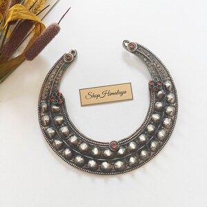 Puede incluir: Un collar de metal plateado en forma de media luna con dos filas de tachuelas cónicas en relieve, patrones grabados e incrustaciones circulares pequeñas en rojo, verde y azul. Una etiqueta rectangular con el texto Shop Himalaya se sitúa en el centro.