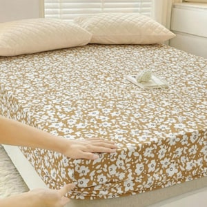 Op de afbeelding: Een hoeslaken met een bloemenpatroon in wit en bruin. Twee beige gewatteerde kussens liggen op het bed. Het laken wordt aangepast door een hand. Het bed staat tegen een raam met jaloezieën. Het laken is ongeveer 200cm x 160cm.