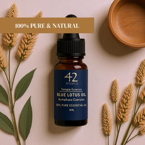 May include: A brown glass bottle of Blue Lotus Oil with a black dropper, set against a pink background. The bottle has a navy blue label with white text that reads "42 Rituals Temple Essence Blue Lotus Oil." The image also includes sprigs of wheat and a wooden bowl. The text "100% Pure & Natural" is displayed.