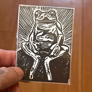 May include: Black and white linocut print of a frog sitting on a rock. The frog is centered, with detailed lines defining its form. The background features radiating lines, creating a sunburst effect. The print is in a clear plastic sleeve.