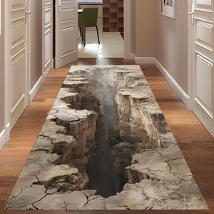 Op de afbeelding: Een rechthoekig vloerkleed met een 3D-canyonontwerp. Het vloerkleed toont een gedetailleerde weergave van een diepe canyon met rotsachtige kliffen en een mistige atmosfeer. Het kleurenpalet omvat tinten bruin, grijs en beige.