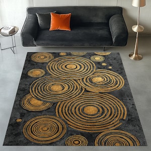 Könnte beinhalten: Rechteckiger Teppich mit einem Muster aus goldenen und schwarzen konzentrischen Kreisen auf dunklem Grauhintergrund. Der Teppich liegt vor einem schwarzen Sofa mit einem orangefarbenen Kissen.