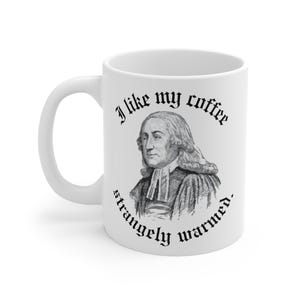 May include: White ceramic coffee mug featuring a black and white illustration of a man with the text "I like my coffee strangely warmed."