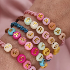 Könnte beinhalten: Eine Sammlung farbenfroher Perlenarmbänder mit Wörtern wie "MAJA", "STRENGTH", "NAMASTE" und "FUCK". Die Perlen sind in verschiedenen Pastellfarben mit goldenen Buchstaben verziert. Die Armbänder befinden sich auf einem rosa Hintergrund.
