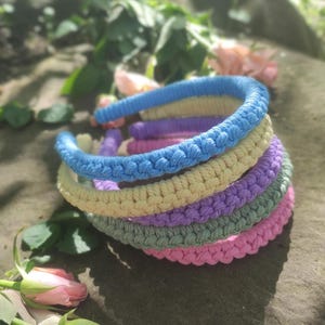 Könnte beinhalten: Ein Stapel bunter Makramee-Haarbänder in Blau, Gelb, Lila, Grün und Rosa. Die Haarbänder bestehen aus gewebtem Kordel und liegen auf einer Steinoberfläche, mit rosa Rosen im Vordergrund.
