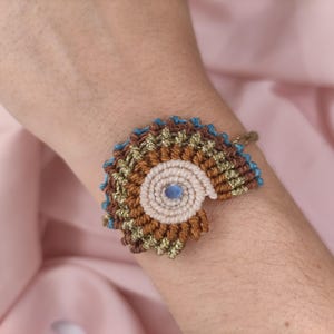 Könnte beinhalten: Ein Makramee-Armband mit einem fächerförmigen Design aus braunen, goldenen und blauen Fäden. Das Armband hat ein zentrales Spiralelement mit einer kleinen blauen Perle. Das Armband wird am Handgelenk getragen.