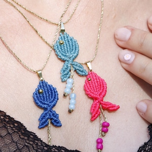 Peut inclure: Trois pendentifs poissons en macramé bleu, bleu clair et rose, suspendus à de fines cordelettes dorées. Les poissons sont formés de fils noués avec des perles métalliques en guise d'yeux et des perles assorties suspendues aux queues.