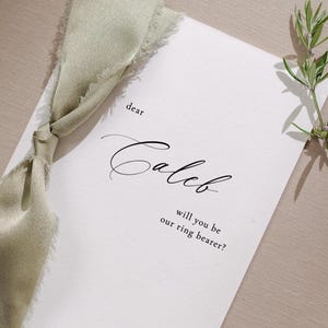 May include: A white card with green ribbon tied around it. The card says "dear Caleb, will you be our ring bearer?"