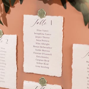 May include: A peach-coloured wedding seating chart with handwritten table assignments. Each table card is fixed with a green wax seal. Floral arrangements of white and peach flowers and greenery adorn the top.