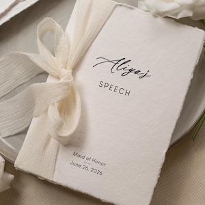 May include: A cream-colored speech booklet tied with a matching ribbon, featuring the name "Aliya's" in elegant script. The word "SPEECH" is printed below, with "Maid of Honor" and "June 26, 2026" at the bottom. The booklet rests on a gray plate.