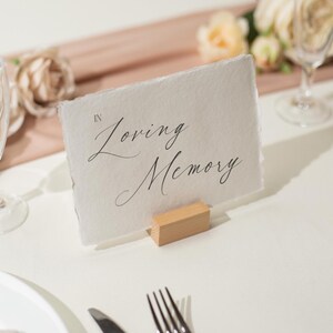 May include: A white card with the words "In Loving Memory" in elegant script, held by a small wooden block. The card sits on a white tablecloth, with champagne flutes and floral arrangements in the background, suggesting a memorial event.