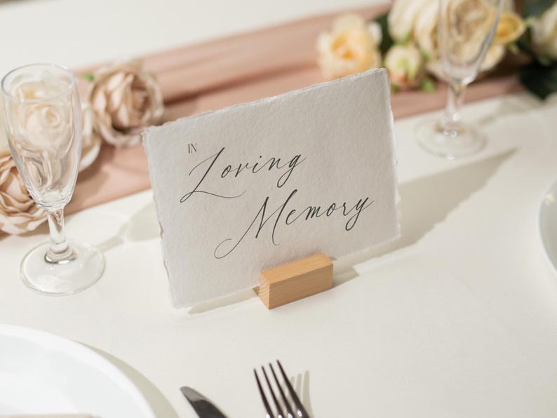 May include: A white card with the words "In Loving Memory" in elegant script, held by a small wooden block. The card sits on a white tablecloth, with champagne flutes and floral arrangements in the background, suggesting a memorial event.