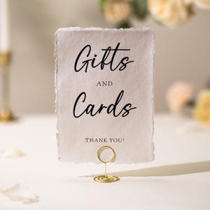 May include: A rectangular sign with the words "Gifts and Cards" in elegant black script, held by a gold wire holder. The sign has a textured, off-white paper appearance and includes the phrase "THANK YOU!"